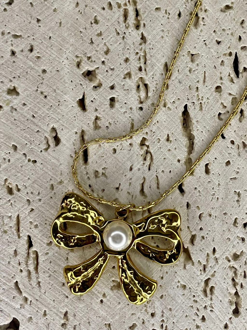 Gold bow-shaped necklace with a pearl center on a textured beige surface