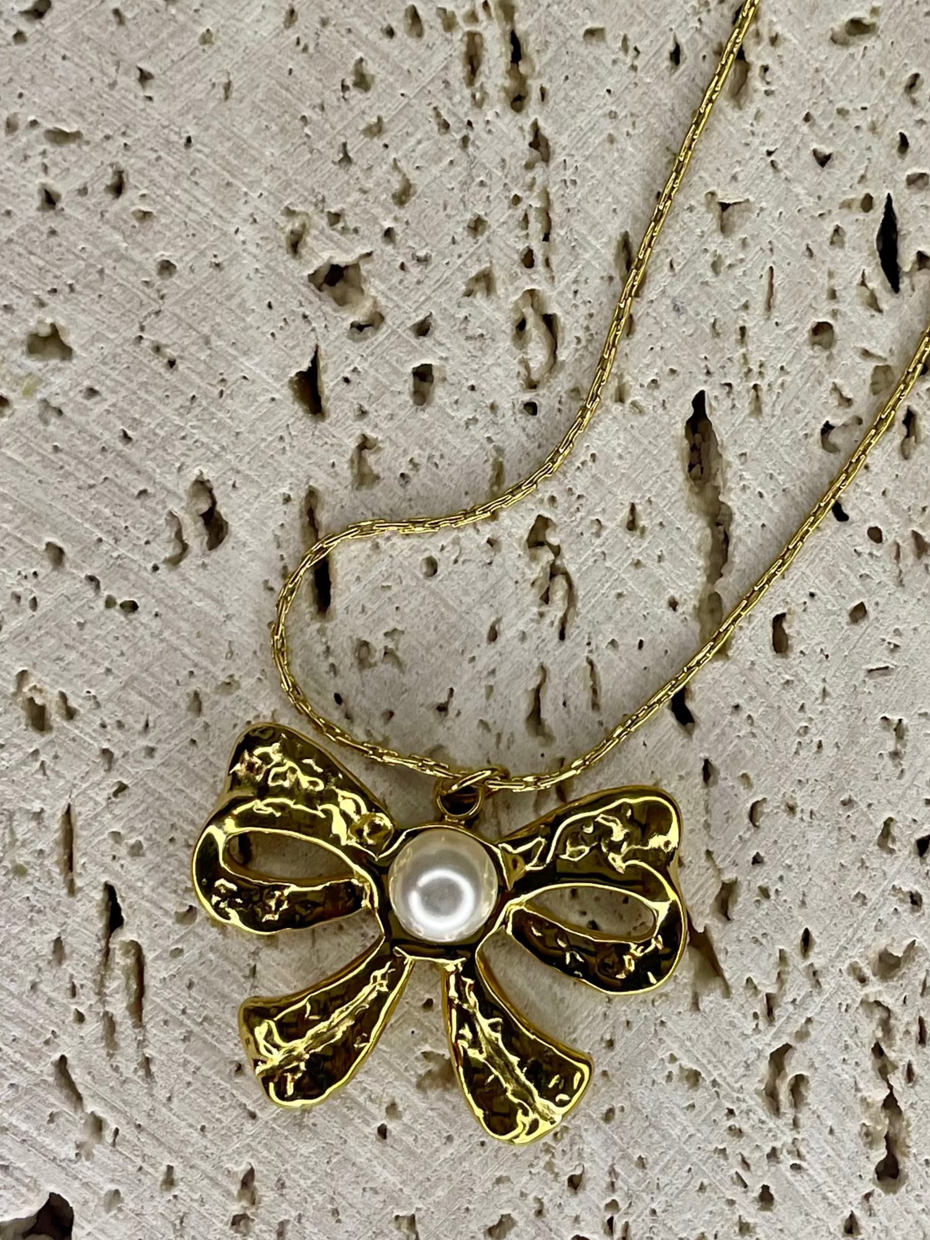 Gold bow-shaped necklace with a pearl center on a textured beige surface